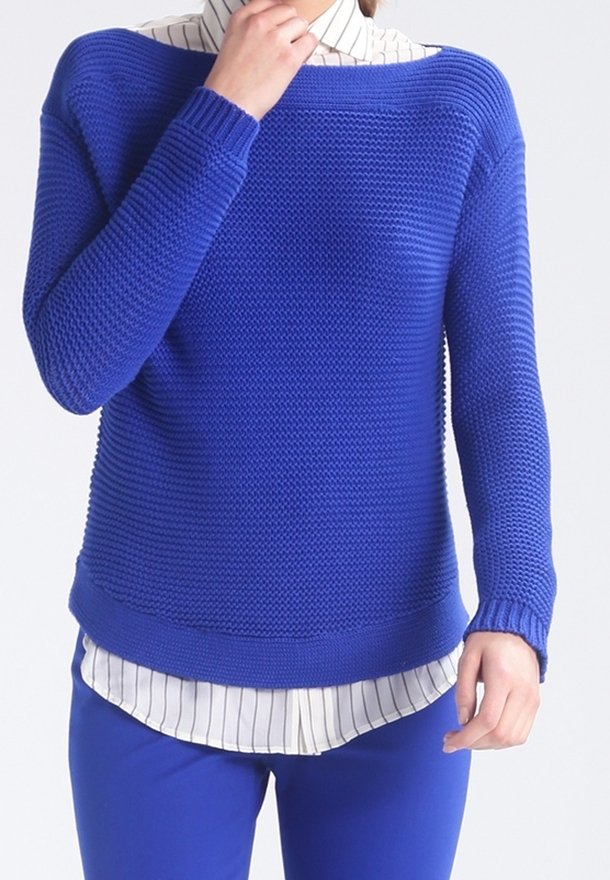 Personne portant un pull en tricot texturé bleu sur une chemise blanche à fines rayures noires et un pantalon bleu assorti, sur un fond uni.