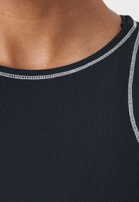 Primer plano de una camiseta sin mangas de color negro con relieve y costuras en blanco, llevada en el cuello y parte superior del pecho de una persona.