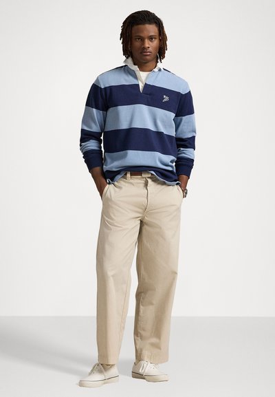 Langärmige rugbyshirt met blauwe en marineblauwe horizontale strepen, witte kraag, gecombineerd met beige chinos en witte sneakers.
