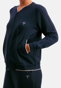 Chaqueta azul marino con cremallera, hecha de tela suave, con un logo discreto, dos bolsillos laterales y ribete en contraste en el cuello y los puños.