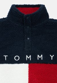 Pile felpato color navy, bianco e rosso con collo alto, tre bottoni a pressione e ricamo "TOMMY" in bianco sul davanti. Texture morbida.