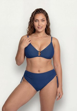 Donna con capelli ricci che indossa un bikini blu con dettagli dorati, in piedi davanti a uno sfondo chiaro uniforme, sorridente verso la macchina fotografica.
