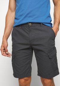Uomo che indossa pantaloni cargo neri con tasche e una maglietta blu, con le mani rilassate ai lati su uno sfondo semplice.