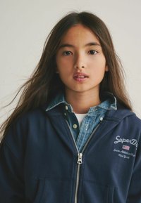 Felpa con zip blu scuro con logo, indossata sopra una camicia di jeans azzurra. Capelli lunghi e castani incorniciano parzialmente il volto, con uno sfondo neutro.