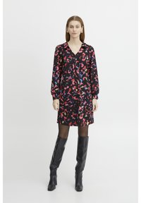 Schwarzes Kleid mit farbenfrohem abstraktem Muster, langen Ärmeln und V-Ausschnitt. Besitzt einen lockeren Schnitt und wird mit schwarzen, knielangen Stiefeln kombiniert.