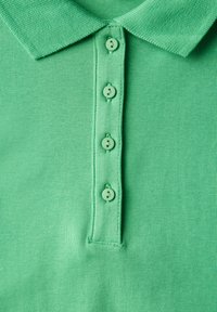 Grön polo-shirt med en texturerad tyg, med en klassisk krage och fem matchande knappar fram. Sömmarna är rena.
