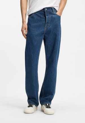 Personne portant un jean bleu droit, mains dans les poches et baskets blanches sur un fond blanc uni.