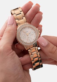 Roségoldene Uhr mit rundem Zifferblatt, Kristallakzenten und Teilzeigern. Das Armband besteht aus verschlungenen Metallgliedern und hat eine polierte Oberfläche.