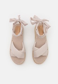 Sandalias de esparto beige con correas de tela cruzadas, lazos atados en el tobillo y una suela envuelta en yute. Textura suave y diseño plano.