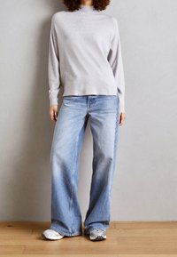 Grå långärmad topp med hög hals, i kombination med ljusblå vida jeans och grå-vita sneakers, på ett trägolv.