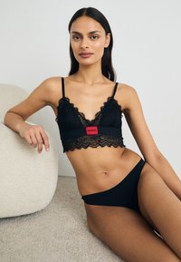Svart blond bøylebh med bølgete kanter, utstyrt med en rød logo-stripe. Kombinert med matchende svarte truser, vist på en lys teksturert overflate.