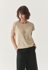 Beige pletený top s krátkymi rukávmi a okrúhlym výstrihom, kombinovaný s čiernymi širokými nohavicami. Hladká textúra, minimalistický dizajn.