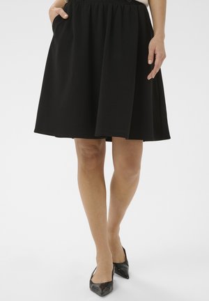 Femme portant une jupe noire évasée longueur genou avec poches et des ballerines noires à bout pointu, debout avec une jambe légèrement avancée.