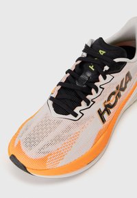 Chaussure de course Hoka en gris avec des accents oranges, dotée d'une tige en maille, de lacets noirs et d'un logo proéminent sur le côté. Semelle en caoutchouc.