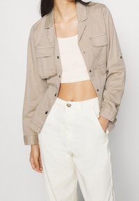 Beige bomuldsjakke med lange ærmer, to brystlommer og sorte knapper, kombineret med en cremefarvet cropped top og hvide bukser med høj talje.