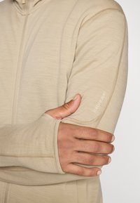 Top bej cu mânecă lungă, dotat cu un fermoar, cu cusături plate și un logo brodat al brandului pe umăr. Textură moale, design ajustat.