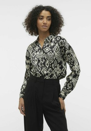 Chemise à motifs noir et blanc avec un col pointu et des manches longues, fabriquée en tissu brillant ; associée à un pantalon noir ajusté.