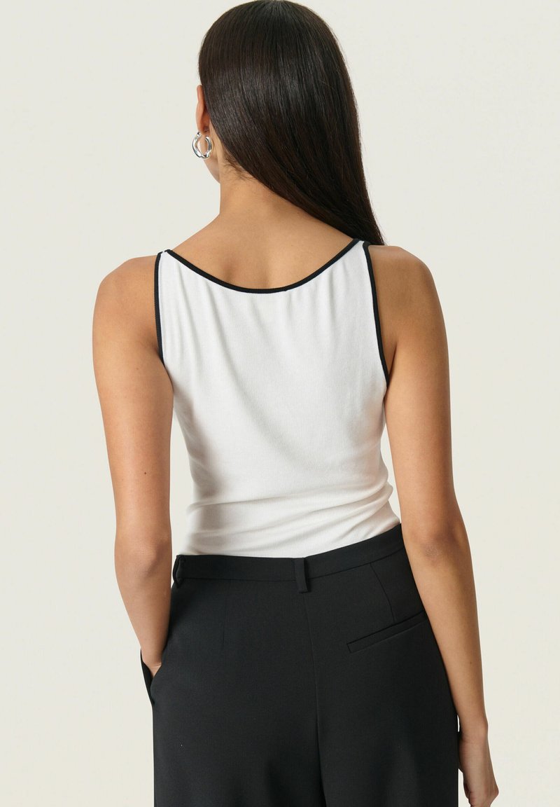 Top blanco sin mangas con un escote en la espalda, ribete negro a lo largo del escote y las sisas, combinado con pantalones negros de cintura alta. Textura de tela suave.
