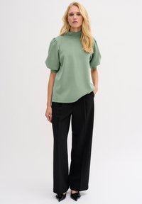 Green short-sleeve top with high collar and gathered shoulders, paired with black wide-leg trousers and pointed-toe shoes.