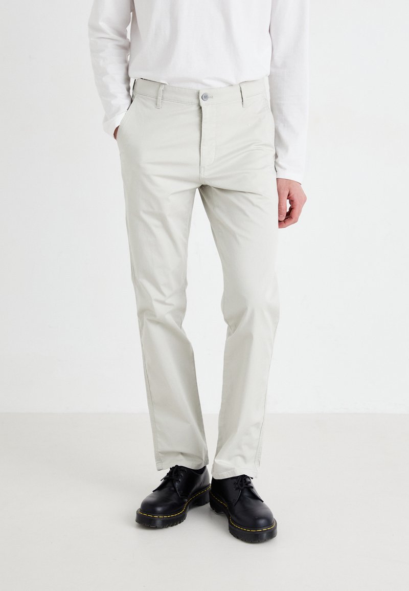 Lys grå, slim-fit bukser laget av en bomullsblandingsstoff, med glidelås og knappelukking, og sidelommer, kombinert med svarte sko.