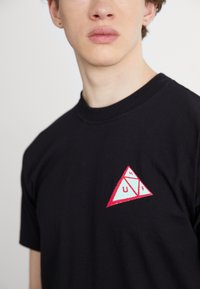 Camiseta de algodón negra con un gráfico en forma de triángulo rojo y azul claro con el texto "HUF". Diseño clásico de cuello redondo y mangas cortas.