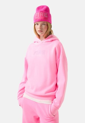 Rosa Kapuzenpullover mit dem Logo "FIRE" in glitzerndem Text. Kombiniert mit passenden Hosen und einer rosa Strickmütze. Weiches, lässiges Material mit lockerer Passform.