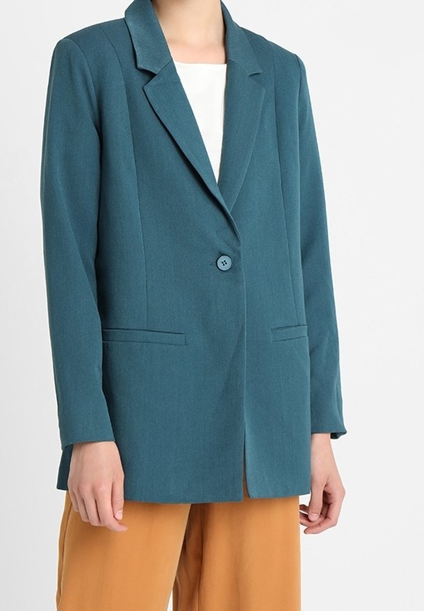 Blazer verde azulado con cuello con muesca, un botón y dos bolsillos frontales. Tela suave y corte entallado, llevado sobre una blusa clara y pantalones marrón claro.