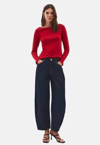 Kvinde med langt brunt hår iført en rød langærmet bluse, vide mørkeblå jeans og sorte højhælede sandaler, stående med hænderne i lommerne.