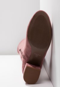 Bottine en daim rose reposant sur la semelle contre des murs blancs, montrant la semelle en caoutchouc brun texturée, le talon bloc, la fermeture éclair latérale et l'étiquette taille 37.