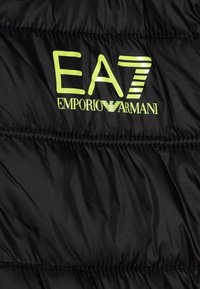 Črna puhovka z sijočo prevleko, ki ima EA7 logotip v svetlo rumeno-zeleni barvi. Strukturirana s horizontalnimi šivi.