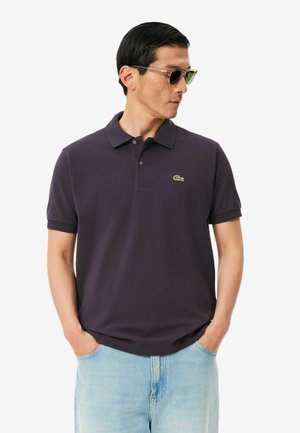 Hombre con camiseta polo morado oscuro con pequeño logo verde de cocodrilo, vaqueros azul claro y gafas de sol, de pie con las manos en los bolsillos.