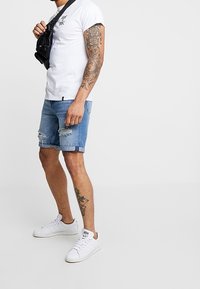 T-shirt blanc à manches courtes, short en denim avec des bords effrangés, baskets blanches. Look décontracté avec un sac bandoulière noir et un tatouage visible sur le bras.