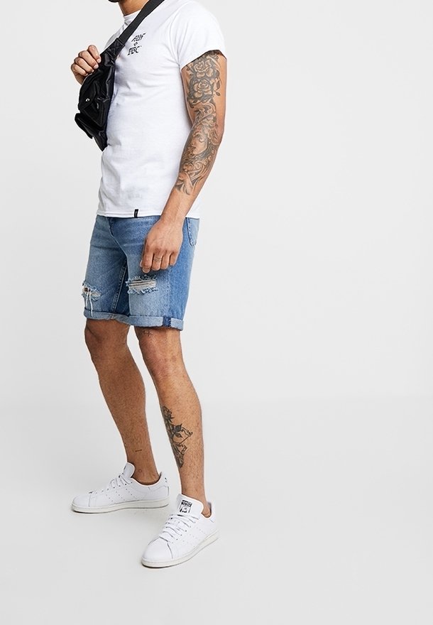 T-shirt blanc à manches courtes, short en denim avec des bords effrangés, baskets blanches. Look décontracté avec un sac bandoulière noir et un tatouage visible sur le bras.