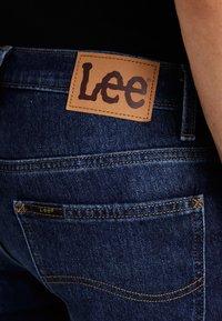 Jeans de mezclilla en azul oscuro con un parche de cuero marrón que presenta el logo "Lee", detalles de costura visibles y una etiqueta más pequeña encima del bolsillo trasero.