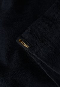 Tela de color azul marino con un tejido texturizado que presenta una etiqueta negra prominente con la inscripción "SUPERDRY" en texto dorado.