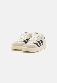 White and cream athletic shoes with black stripes, made of leather and suede. Features gold hardware accents and textured rubber sole.