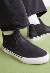 Chaussures noires à enfiler avec une tige en cuir texturé, semelle en caoutchouc blanche et rayures blanches contrastantes le long du bord, portées avec des chaussettes montantes.