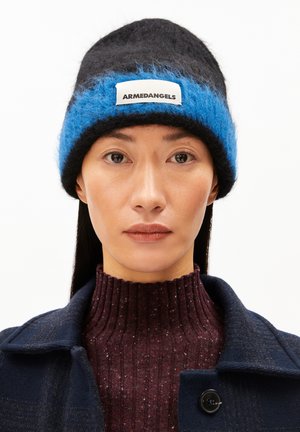 Schwarze und blaue gestrickte Mütze mit einem flauschigen blauen Streifen und einem weißen Etikett. Getragen über einem gerippten kastanienbraunen Pullover und kombiniert mit einem marineblauen Mantel.