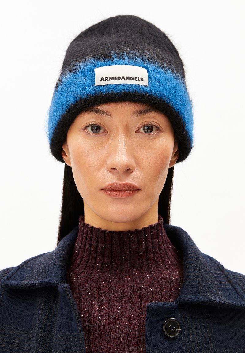 Zwarte en blauwe gebreide beanie met een fuzzy blauwe streep en een wit label. Draagt over een geribbelde bordeauxrode trui en gelaagd met een marineblauwe jas.