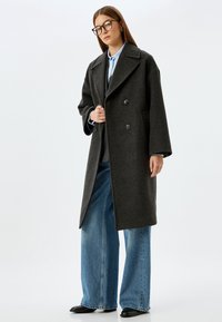 Donkergrijze wollen jas met een dubbele knoopsluiting, grote revers en zijzakken, gecombineerd met blauwe jeans met wijde pijpen en zwarte schoenen.