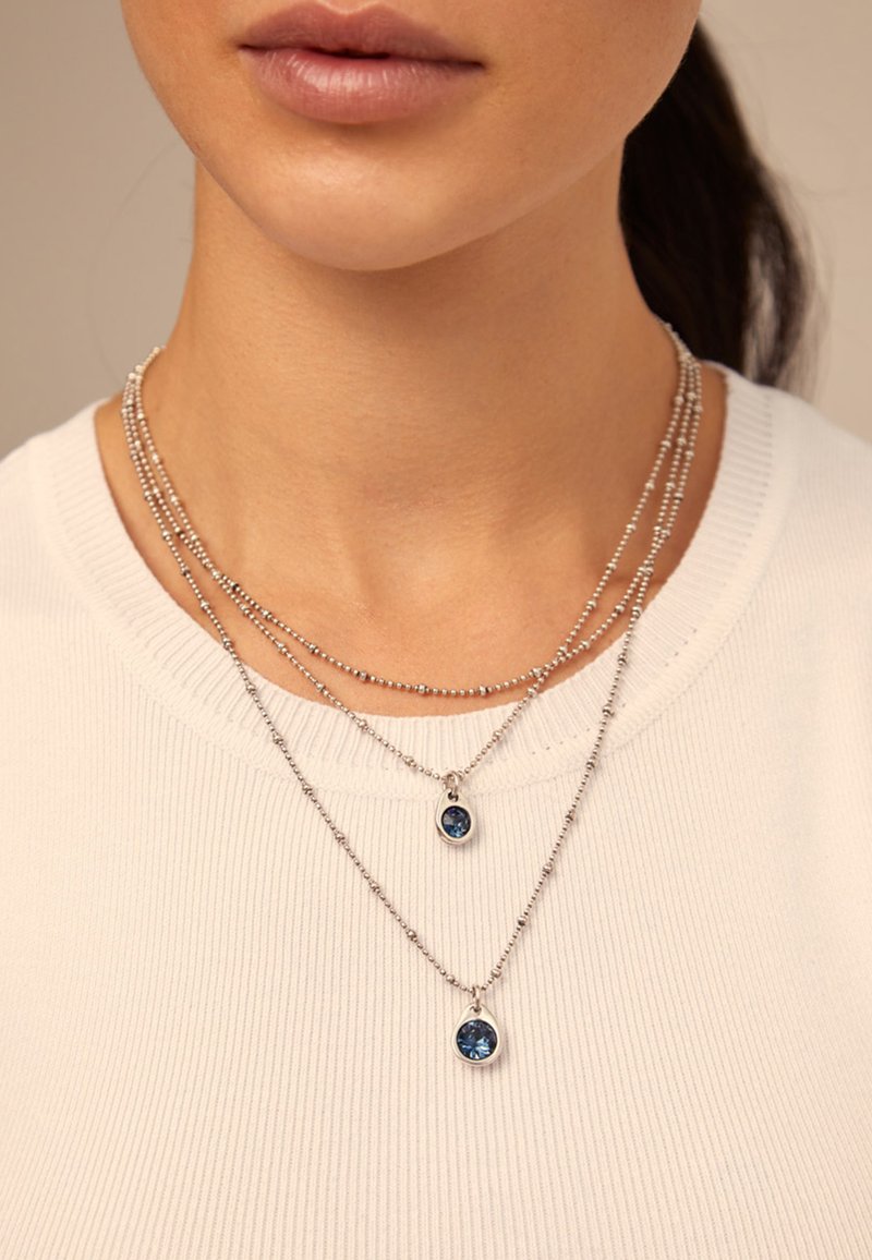 Gros plan sur des colliers en chaîne à billes en argent superposés avec des pendentifs en pierre précieuse bleue, portés par une personne vêtue d'un t-shirt à rayures blanches.