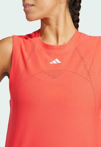 Czerwona bezrękawnik sportowy wykonany z gładkiego materiału, z okrągłym dekoltem, perforowanymi detalami i małym białym logo na klatce piersiowej.