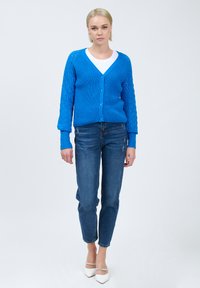 Blauer gestrickter Cardigan mit strukturiertem Muster, Knopfverschluss und V-Ausschnitt. Kombiniert mit enganliegenden Jeans und weißen High Heels.