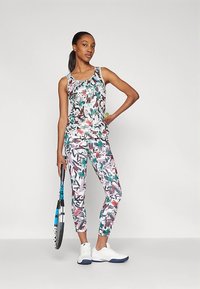 Komplet aktivne obleke, ki vključuje brezrokavno belo majico z barvnim vzorcem in usklajenimi legicami; oba sta izdelana iz raztegljive tkanine. Oseba drži teniški lopar.