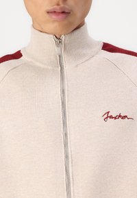 Béžový sveter na zips s vysokým golierom, so sakramentskými akcentmi na pleciach v burgundskej farbe a červeným vyšívaným textom. Hladká textura s pleteným povrchom.