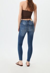 Mujer de espaldas vestida con jeans ajustados azules, una blusa marrón sin mangas y sandalias marrones sobre un fondo blanco liso.