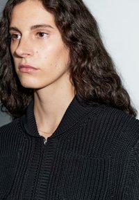 Black zip-up jacket with a textured knit design, featuring a ribbed collar. Worn over a delicate gold chain necklace.