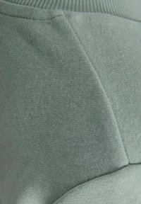 Világoszöld textil, amely sima felülettel és diszkrét heather mintával rendelkezik, varrásokkal, amelyek dimenzionális panelek kialakítását segítik elő, sportos formát adva.