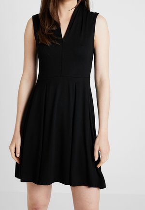 Femme portant une robe noire sans manches à encolure en V avec une jupe évasée, debout devant un fond gris uni.