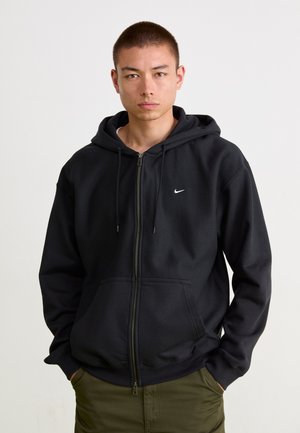 Juodas užtrauktukas džemperis pagamintas iš medvilnės mišinio, su priekinėmis kišenėmis, kapišonu su raišteliais ir mažuoju baltu "Nike" logotipu ant krūtinės.